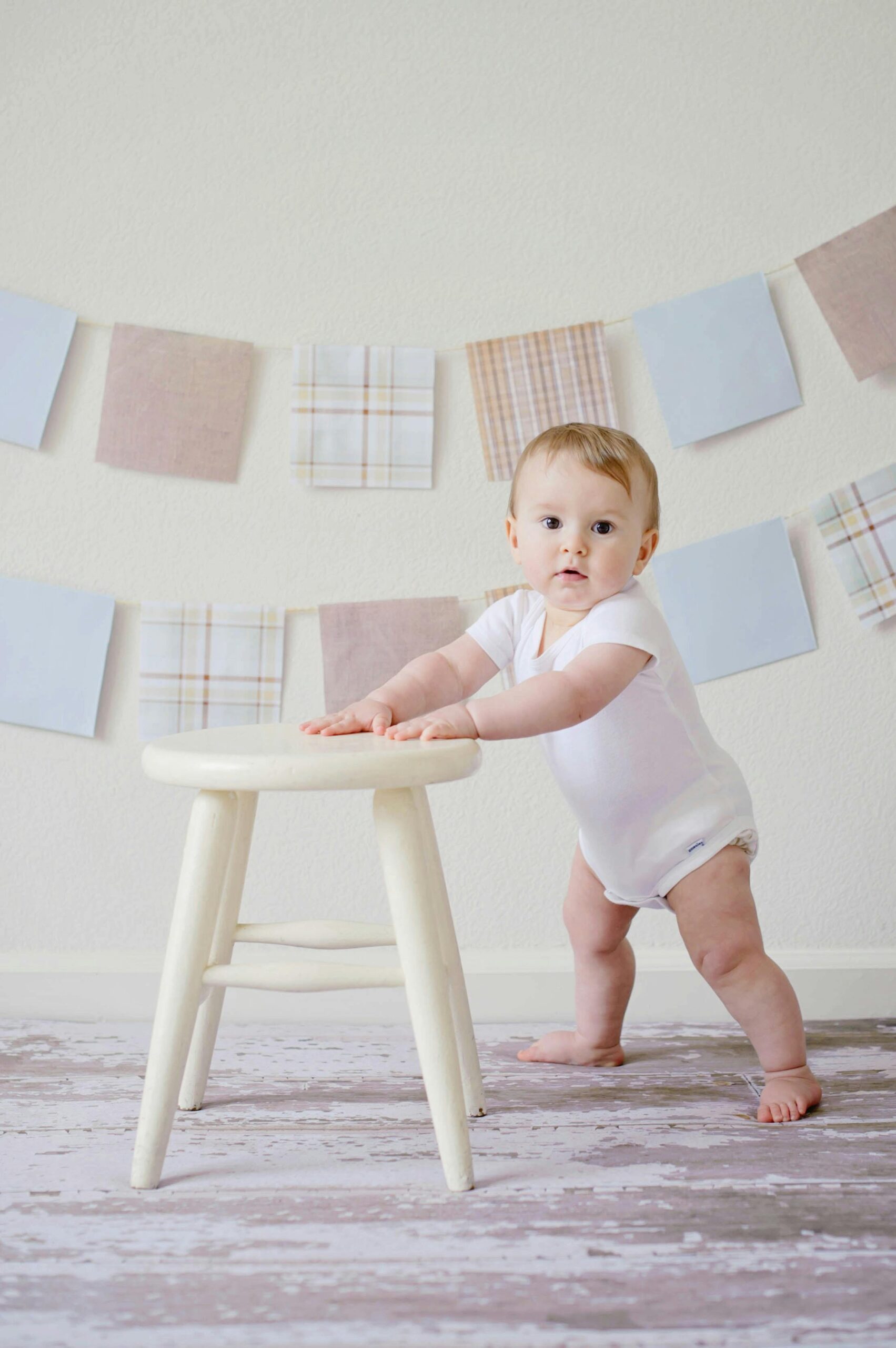 Cute baby standing by a stool indoors with a playful background.