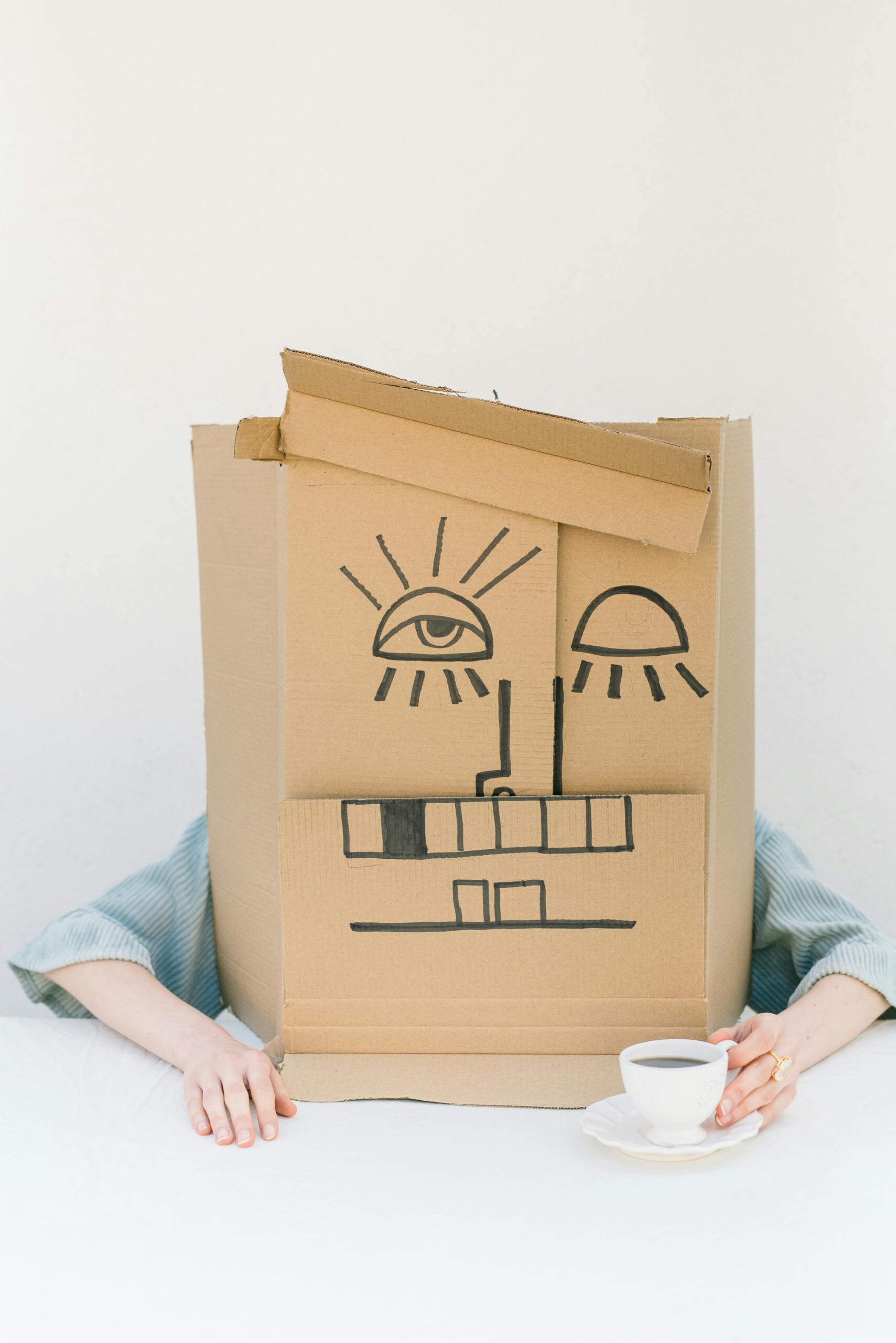 Person wearing a cardboard box mask, sipping coffee at a minimalist setup.